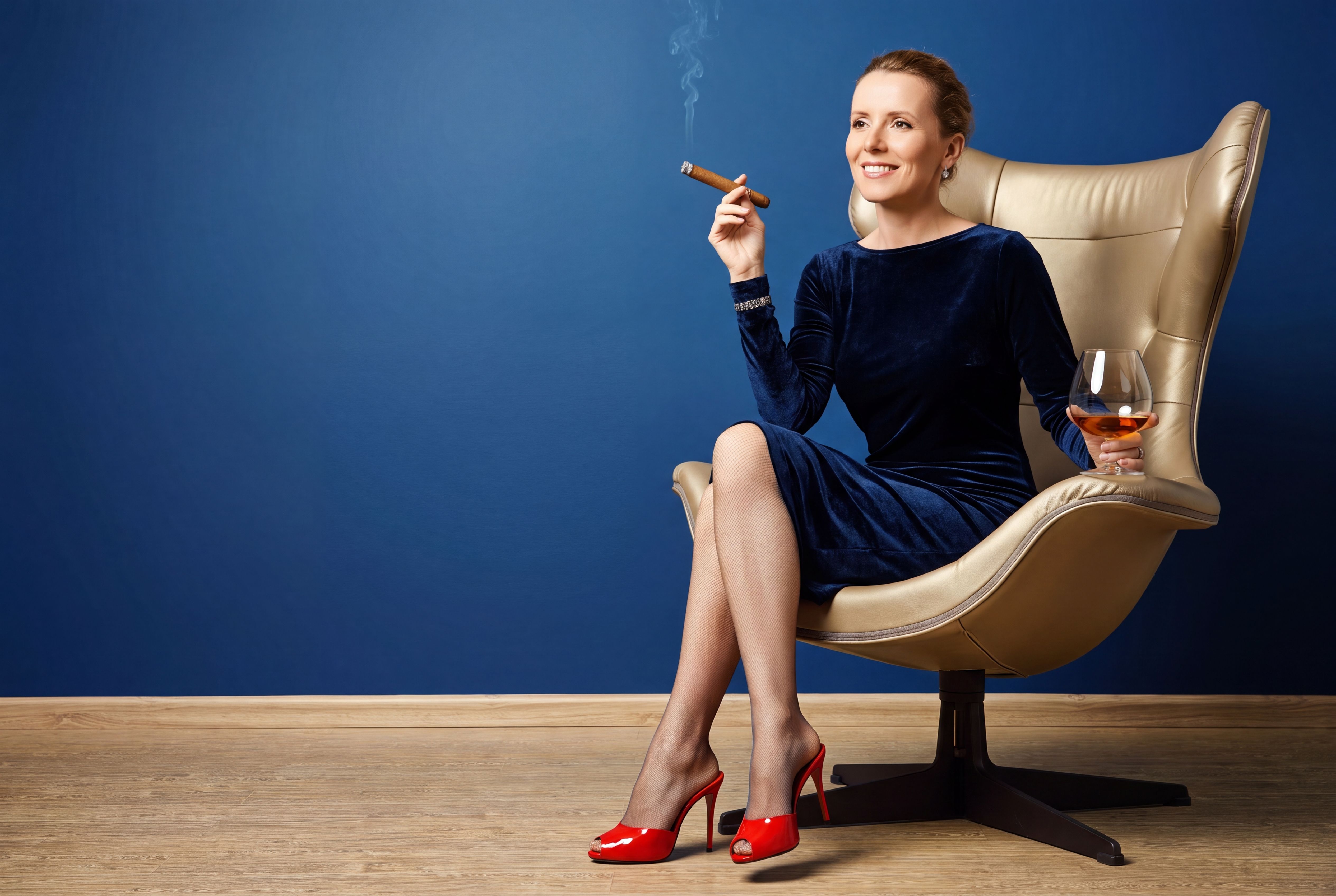 A woman at ease in a sculptural gold chair against deep blue, a cigar in one hand and a glass of cognac in the other, red heels.